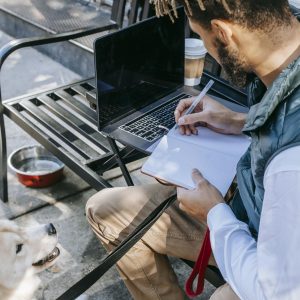 Man taking notes on notepad with a dog by his side; working remotely in an urban setting.