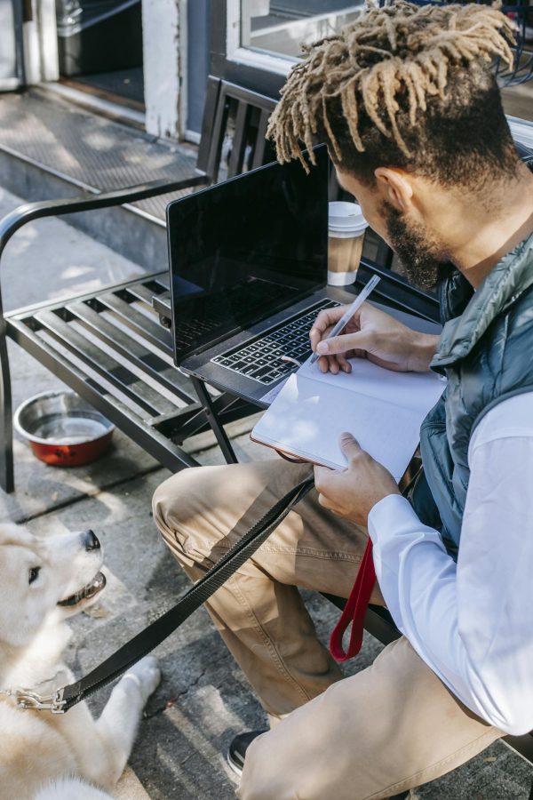 Man taking notes on notepad with a dog by his side; working remotely in an urban setting.